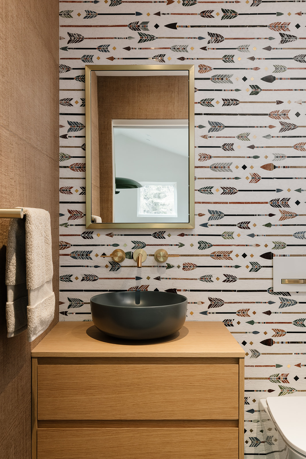 Powder room with a floating oak vanity, arrow-patterned mosaic backsplash, patterned wallcovering, and brushed gold fixtures.