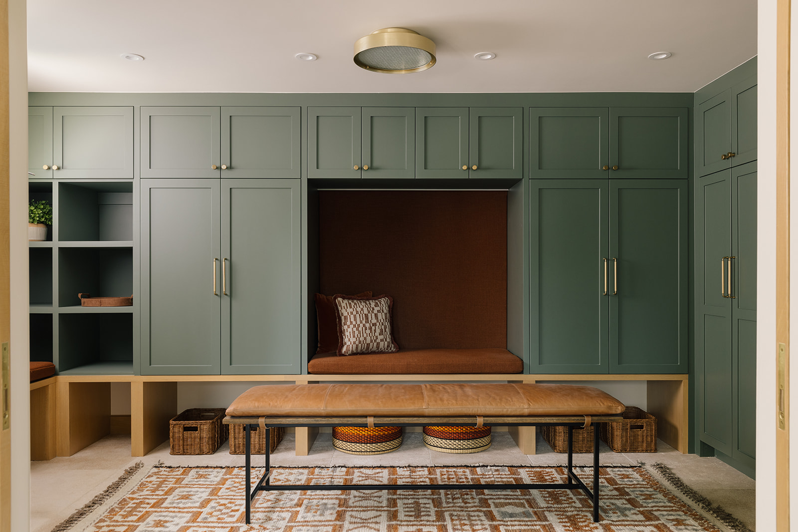 Mudroom with deep green cabinetry, built-in seating with rust-colored upholstery and a padded backrest, a freestanding wood bench with a black metal frame, and a patterned rug.